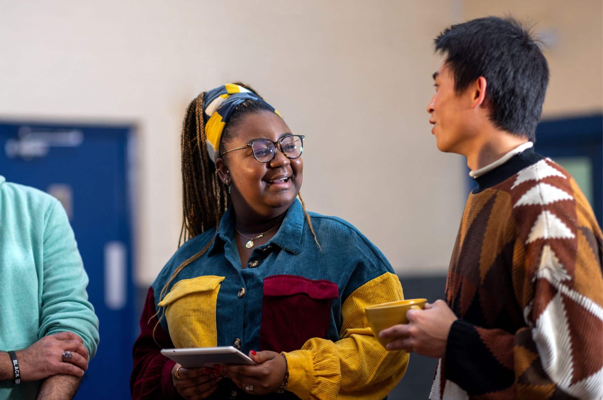 A woman is holding a tablet and smiling whilst talking to a man. The man is holding a coffee cup. They're standing in a community centre.