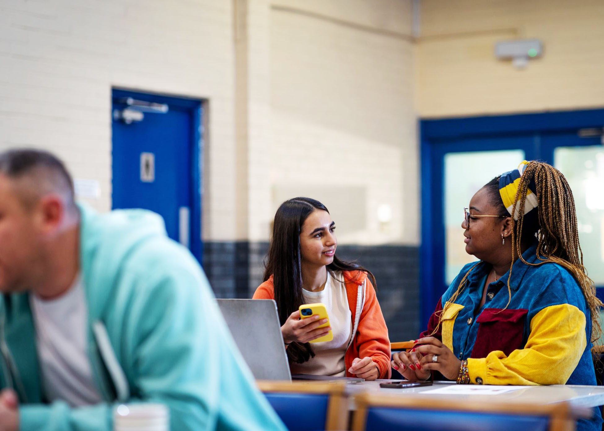 Two people are sat at a table talking in a community hub, one holding a mobile phone with a laptop in front of them