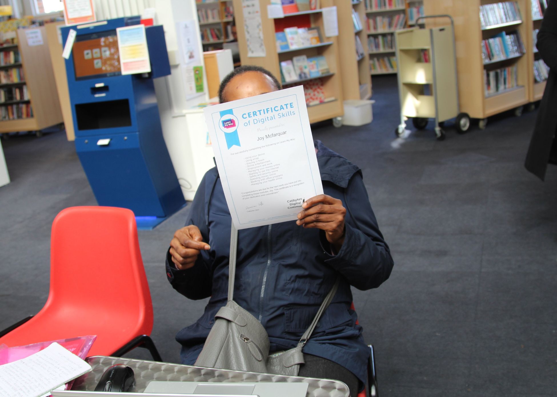 Joy holding a certificate of completion for Learn My Way, sat in a library