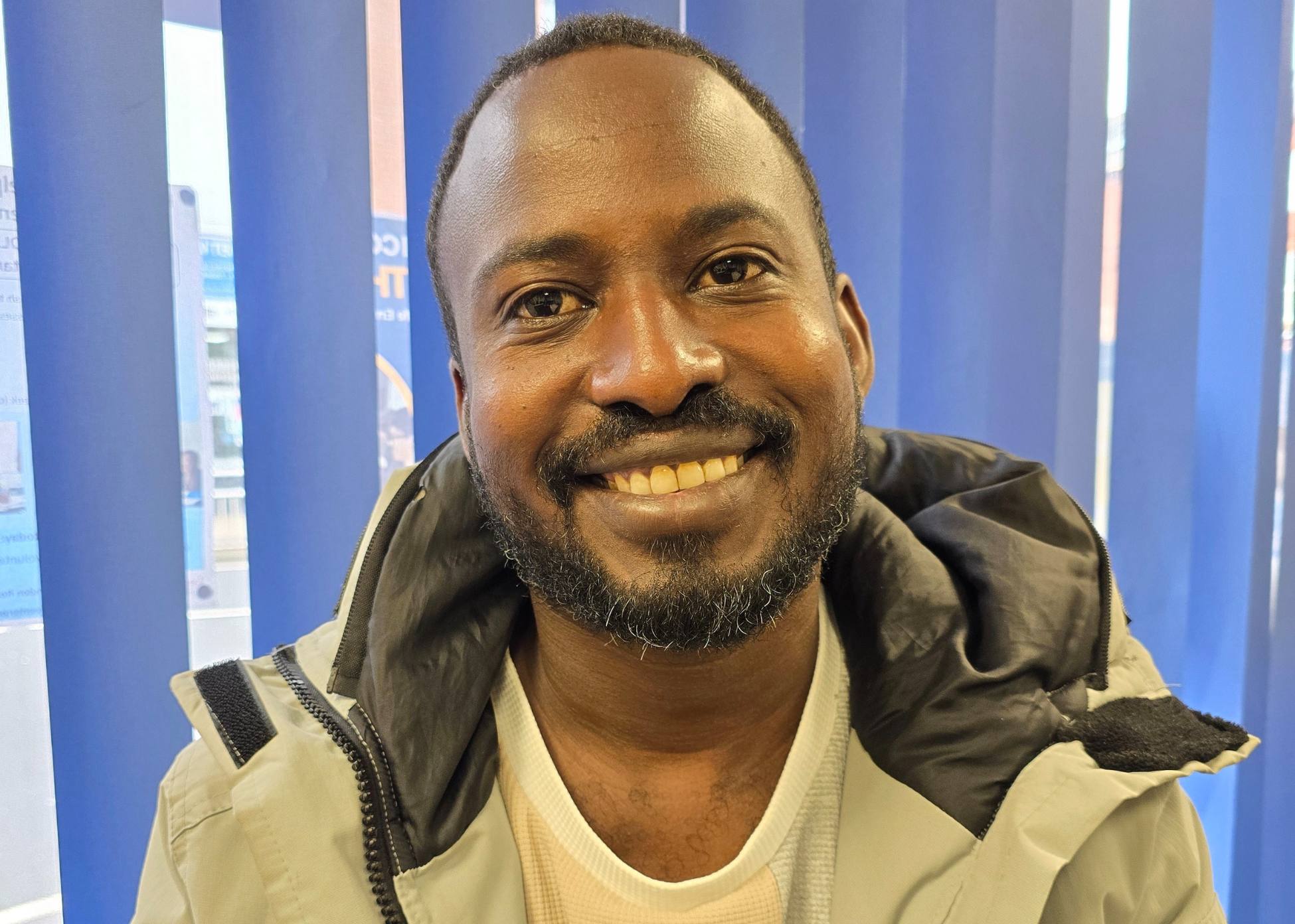 A person looking towards the camera and smiling, wearing a white t-shirt and a coat.