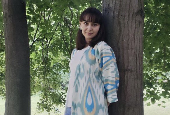 Individual, Madina, leaning against the trunk of a tree, smiling at the camera